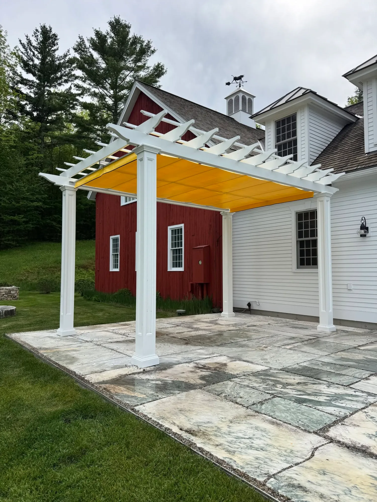 Pergola with shade canopy over a stone patio between a house and a barn